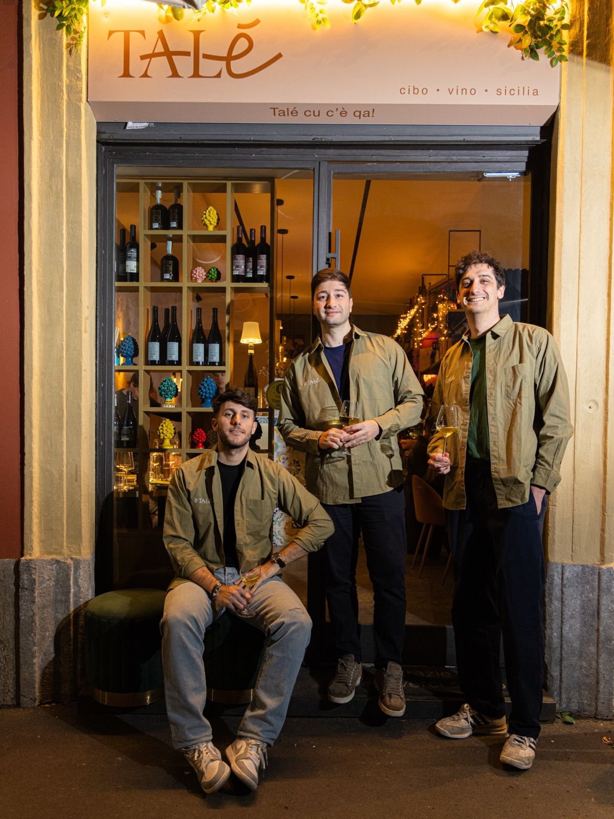 I titolari di Talé Milano Isola davanti all'ingresso del ristorante in Via Pastrengo, con calici di vino e decorazioni di pigne siciliane in vetrina.