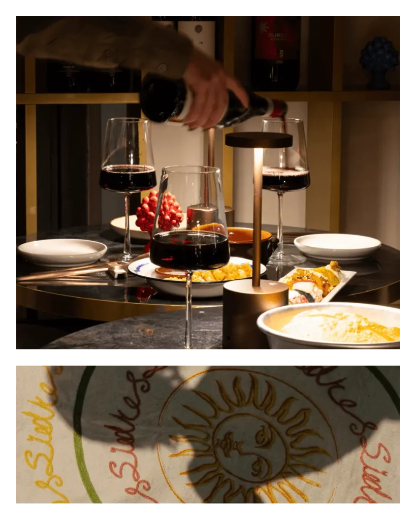 Servizio del vino da Talé Milano Isola: una mano versa vino rosso in un calice su un tavolo imbandito con piatti gourmet e grafica del sole siciliano.